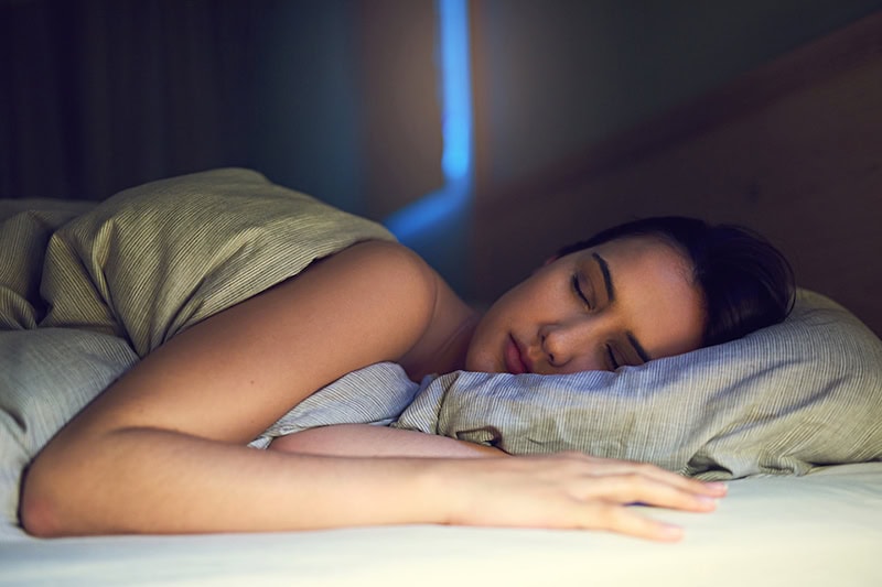 3 Health Benefits for Using Your AC While You Sleep. A young woman sound asleep in her bedroom.