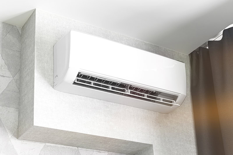 A modern, sleek white mini-split air conditioner mounted near the ceiling on a textured grey wall section, with its air louver slightly open.