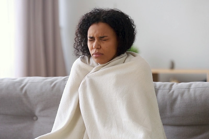 Video - Upgrade Your Furnace. Image is a photograph of a woman sitting on the couch wrapped in a blanket and looking cold.